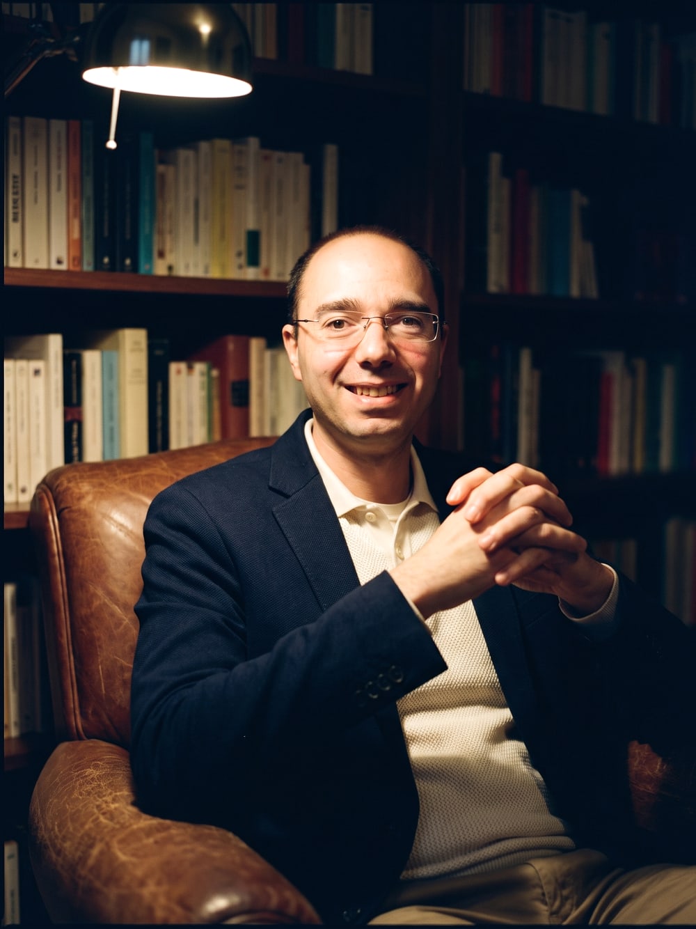 Carlos Hering sentado numa poltrona diante de estantes de livros, sorrindo, em luz quente de abajur.